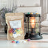 fruity cereal wax melt bag next to lit burner. there are wax melts on the table scattered about to shoe the pieces - these are cute cereal pieces in all the colors along with white "milk" looking wax brittles