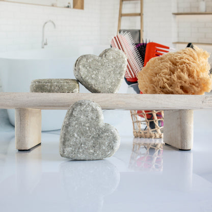 3 green colored green clean shampoo bars shaped in a heart. One is leaning on its side while the other two are on a trey, one leaning on the other. there is a loofah next to them with a cup of hair tools in the background