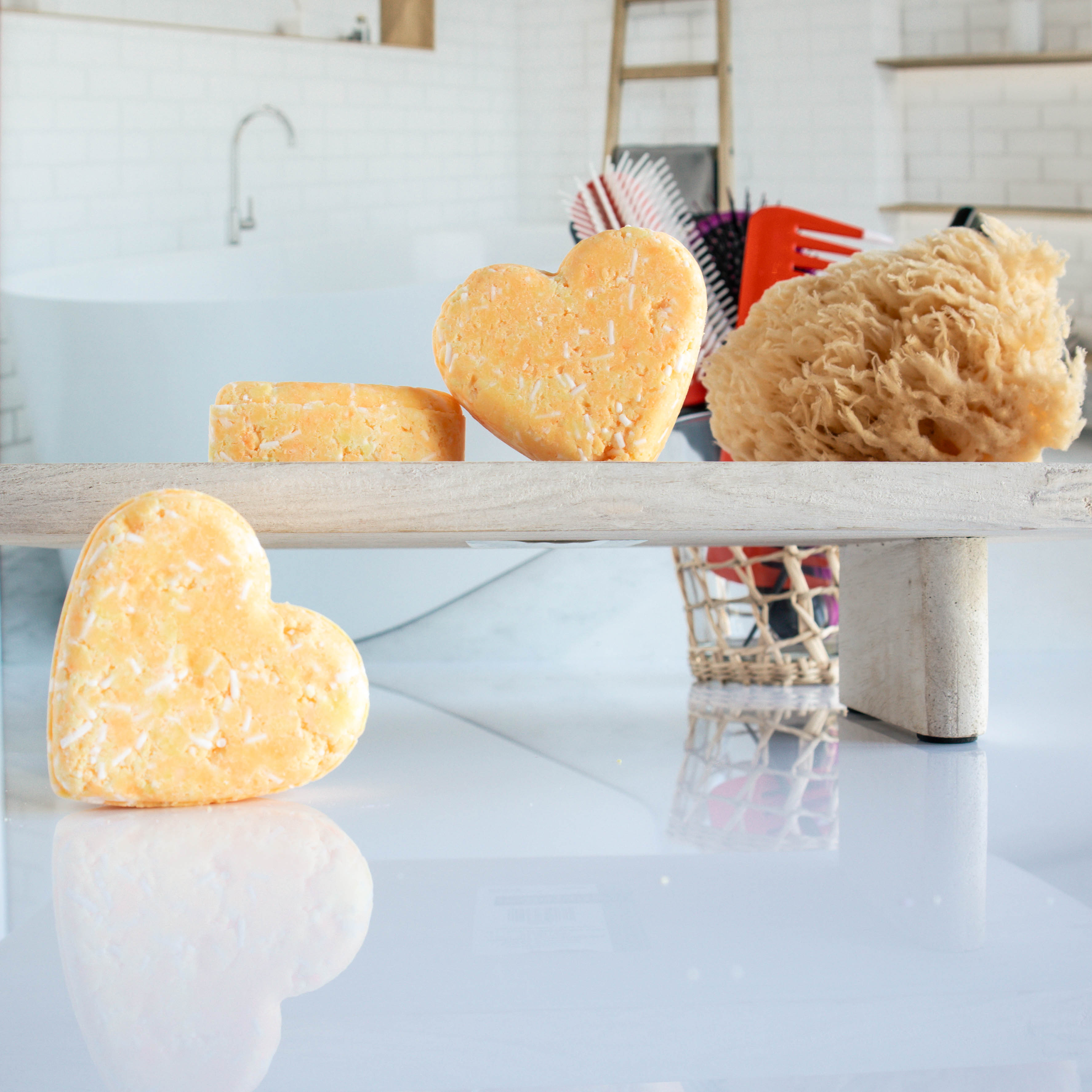 3 light orange harmony shampoo bars shaped in a heart. One is leaning on its side while the other two are on a trey, one leaning on the other. there is a loofah next to them with a cup of hair tools in the background