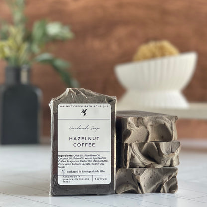 hazelnut coffee soaps are shown, one standing facing out to show the label. Next to this are three more soaps stacked on top to show the sculpted tops. in the background is a soap dish on a stand and a small vase with some greenery