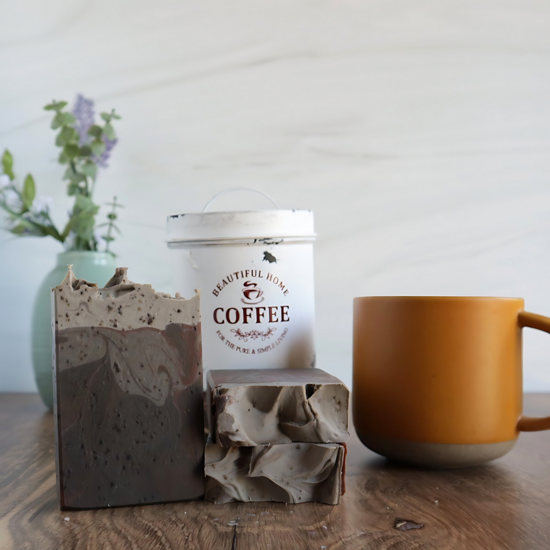 3 hazelnut coffee soaps are shown, one standing facing you to show the dark to light swirls and specs of real coffee grounds throughout.  2 more are laying flat to show a sculpted top. to the right of these is a coral colored coffee cup and in the background there is an antique white tin with the words coffee on it.