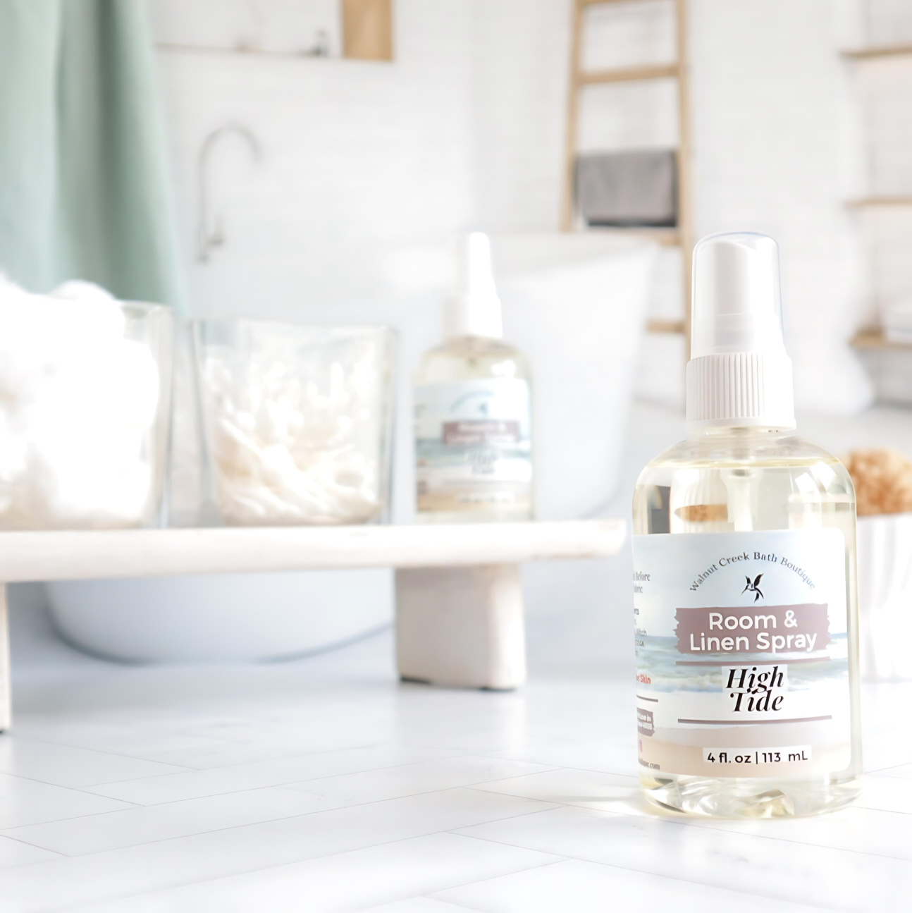 high tide room and linen spray is front right, behind this is a try with cotton balls and qtips in glass jars with another room spray next to them. in the background is an image of a tub and shelf in a spa like bathroom