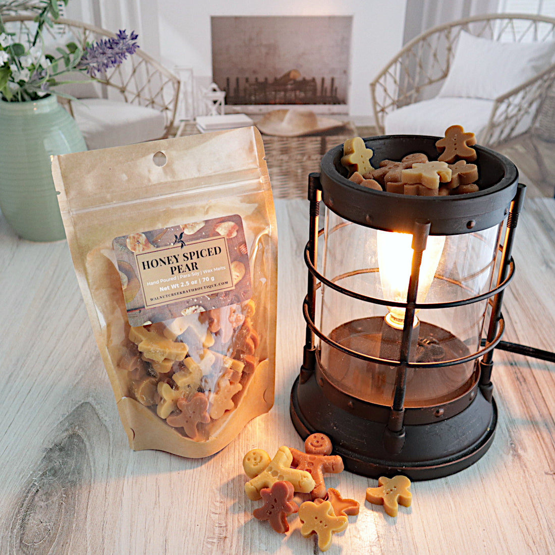 a bag of honey spiced pear wax melts are standing next to a lit burner full of the wax pieces. there are more wax pieces piled next to the burner. the wax is in the shape of small and large gingerbread men, and some are a tan color and some are a light brown color. this is all sitting on a washed out wooden base with a couple of chairs in the background next to a fireplace