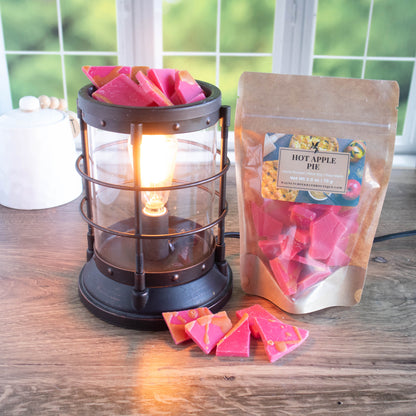 hot apple pie wax melts are red in color with a tan drizzle to represent the pie in the scent. there is a wax burner to the left that is lit with a bowl full of these wax pieces just waiting to be melted! Next to that is a full bag of the wax pieces along with some wax pieces scattered around the base. In the background is a window looking out into a yard . Sitting in front of that is a white crock.