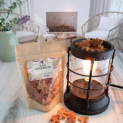 a bag of iced gingersnap wax melts is standing next to a lit burner that is full of the wax pieces. there is a scattering of the wax pieces on the table in front of the burner. the wax is in the shape of light and dark brown gingerbread men. this is all sitting on a washed out wooden base with a couple of chairs in the background next to a fireplace