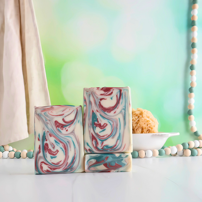 3 icy peppermint soaps are shown with their white base and green and red swirls. one of the soaps is standing on another that is laying flat to show the top of the soap.
there is a soap dish holding a loofah in the background along with some green, white and tan beads. 