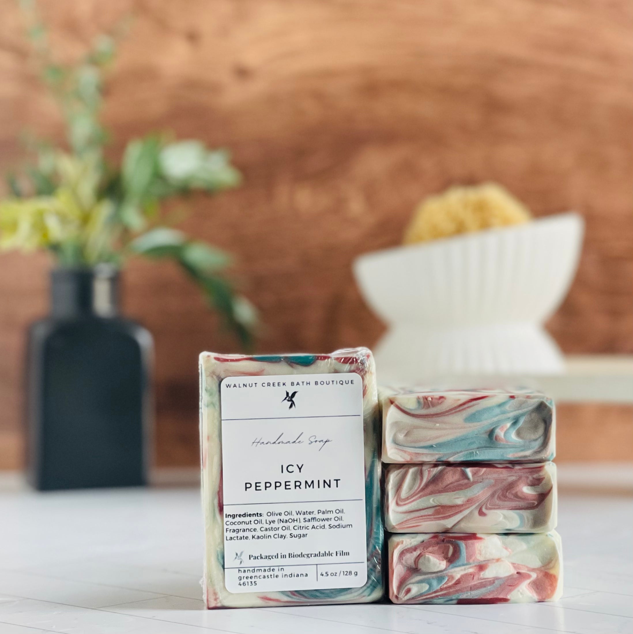 icy peppermint soaps are shown, one standing facing out to show the label. Next to this are three more soaps stacked on top to show the pretty swirls. in the background is a soap dish on a stand and a small vase with some greenery.