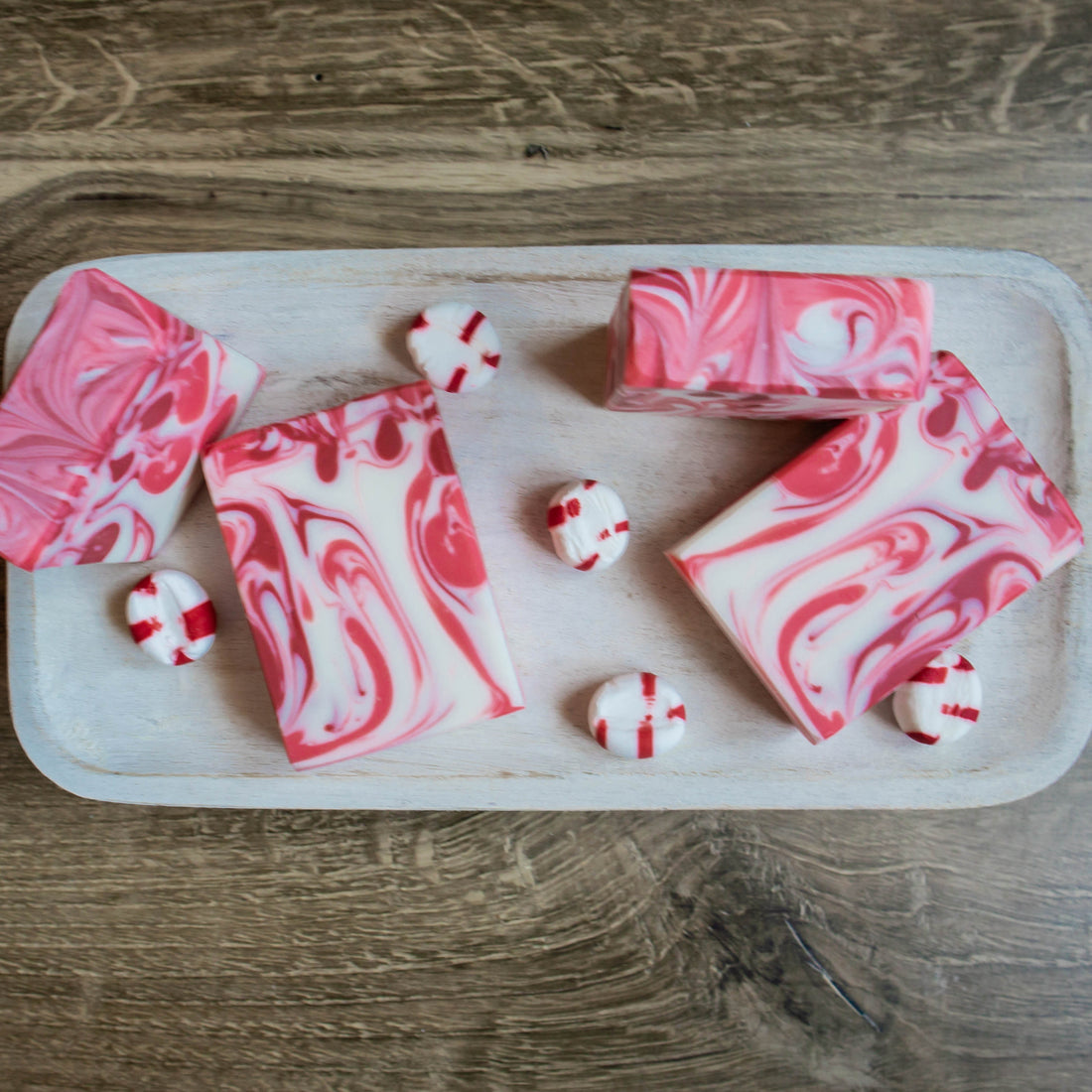 icy peppermint soaps are sitting on a light colored wood tray. 2 are laying flat showing red and pink swirls, two are standing tall showing the pretty swirled tops. there are also some hard peppermint candy scattered around the soaps