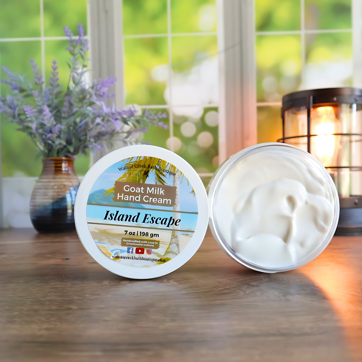 2 island escape creams are shown standing on their sides. One to show the label and the other to show the creamy swirly contents. in the back left is a vase with lavender buds and back right is a lit wax burner to add some light. behind all of this is a window with some greenery showing through.