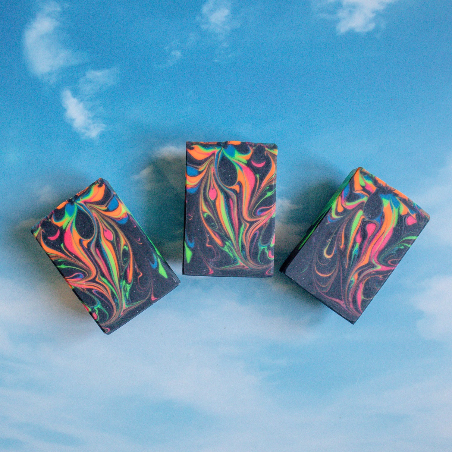a top down view of Jade soap. this beautiful soap is black with neon swirls in the colors of pink, green, blue and orange. they are fanned out and sitting on a blue sky background