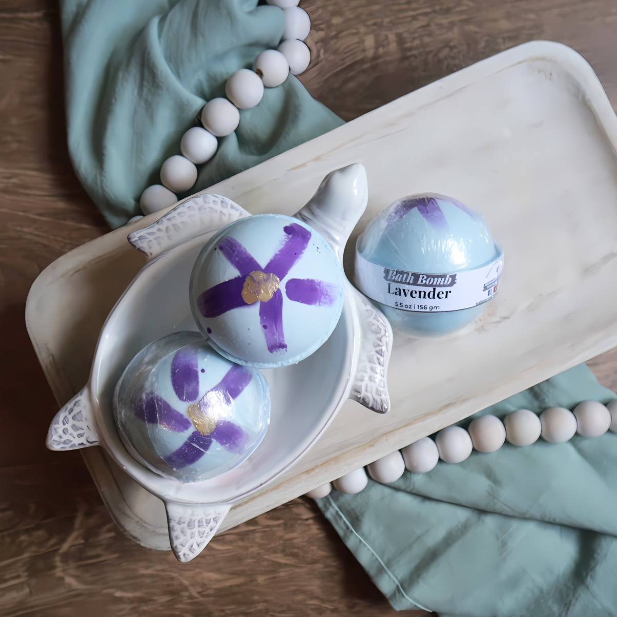 3 lavender bath bombs are shown sitting on a tray. two are in a soap dish in the shape of a turtle. the baht bombs are blue in color with purple swashes along with a gold dap in the middle to represent flower petals. there is a warm wood base and a green towel with white beads running under the tray and through the picture.