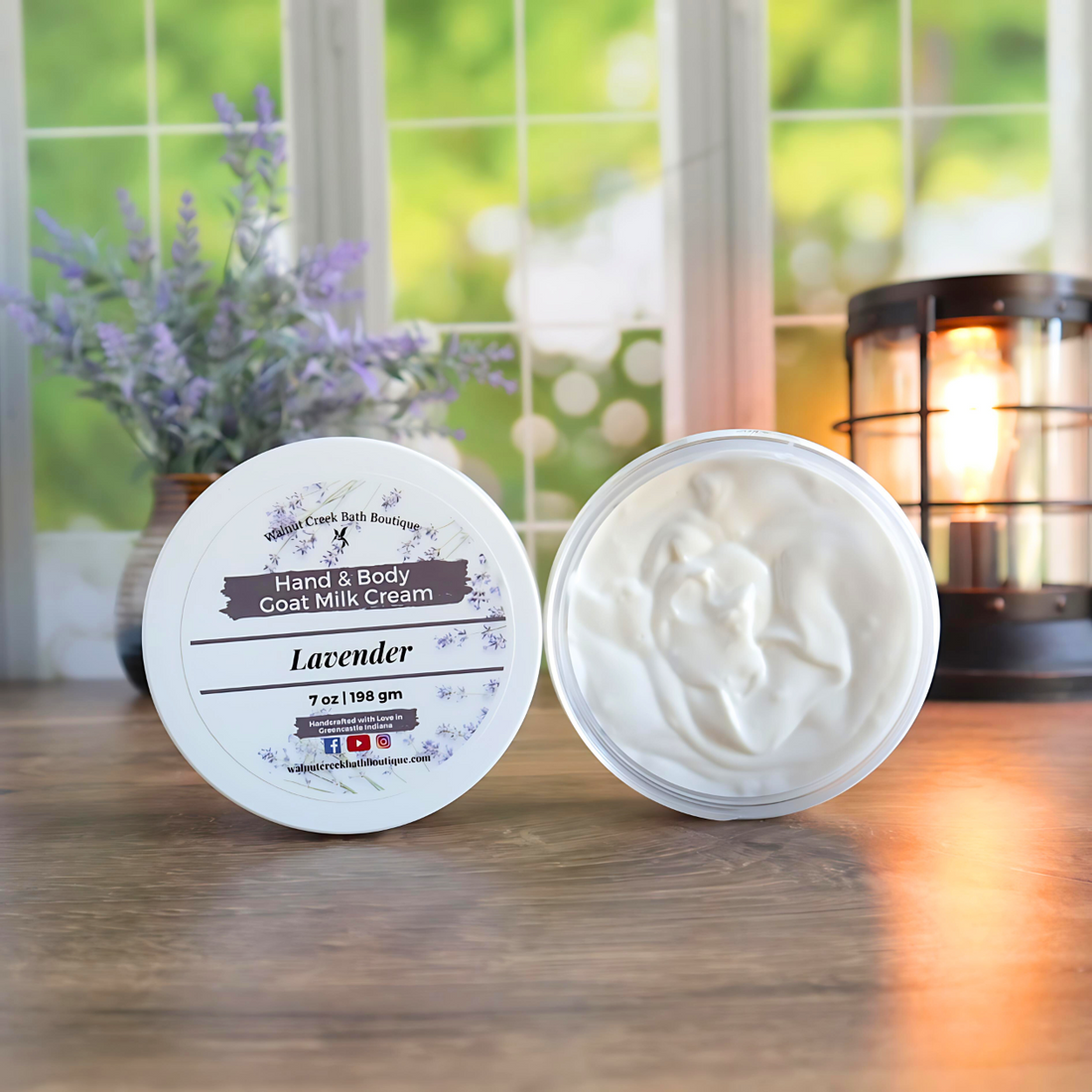 2 lavender creams are shown standing on their sides. One to show the label and the other to show the creamy swirly contents. in the back left is a vase with lavender buds and back right is a lit wax burner to add some light. behind all of this is a window with some greenery showing through.