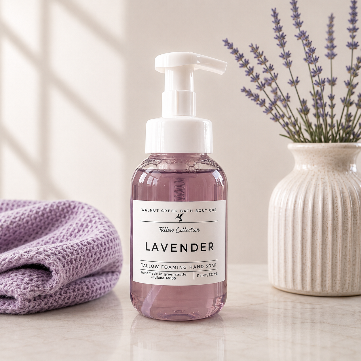 A bottle of Walnut Creek Bath Boutique Lavender Fallow Foaming Hand Soap stands on a light marble surface, flanked by a folded lavender towel on the left and a ceramic vase with lavender sprigs on the right.