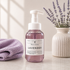 A bottle of Walnut Creek Bath Boutique Lavender Fallow Foaming Hand Soap stands on a light marble surface, flanked by a folded lavender towel on the left and a ceramic vase with lavender sprigs on the right.