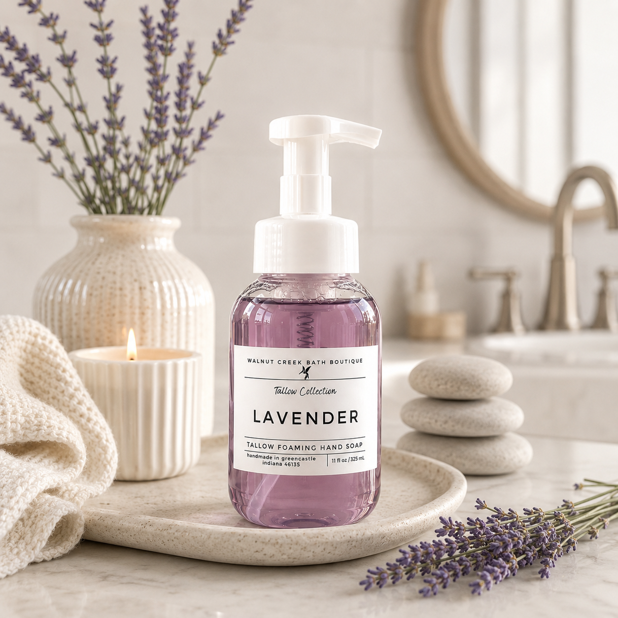 A bottle of Walnut Creek Bath &amp; Body Co. Lavender Tallow Foaming Hand Soap sits on a stone tray in a bathroom setting, surrounded by lavender sprigs, a lit candle, stacked bath stones, and a textured vase.