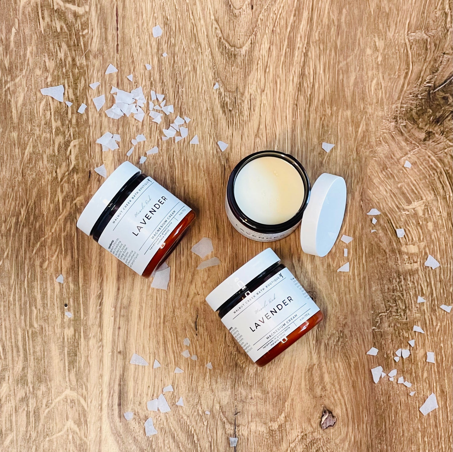 2 jars of magnesium cream are laying flat on a wooden surface with magnesium salt flakes scattered around. there is a 3rd jar sitting upright with the lid off to show the creamy texture of the cream