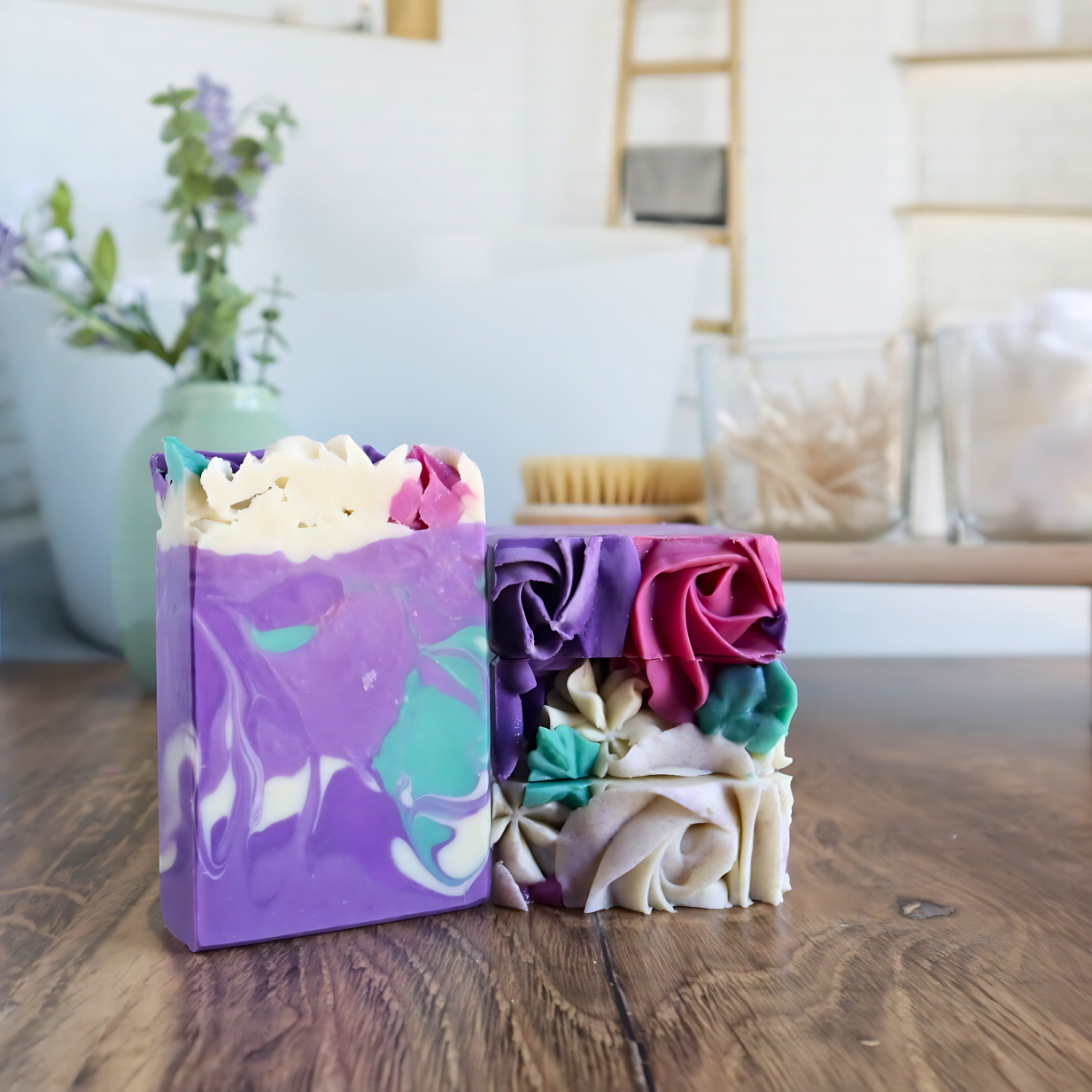 3 lavender soaps are shown, one standing facing you to show the purple base with a darker purple swirl along with white and green swirls. 2 more are laying flat to show the piped top of purple, rose, white flowers and green leaves.  the background is a bath room with a tray of q-tips and cotton balls in a jar.