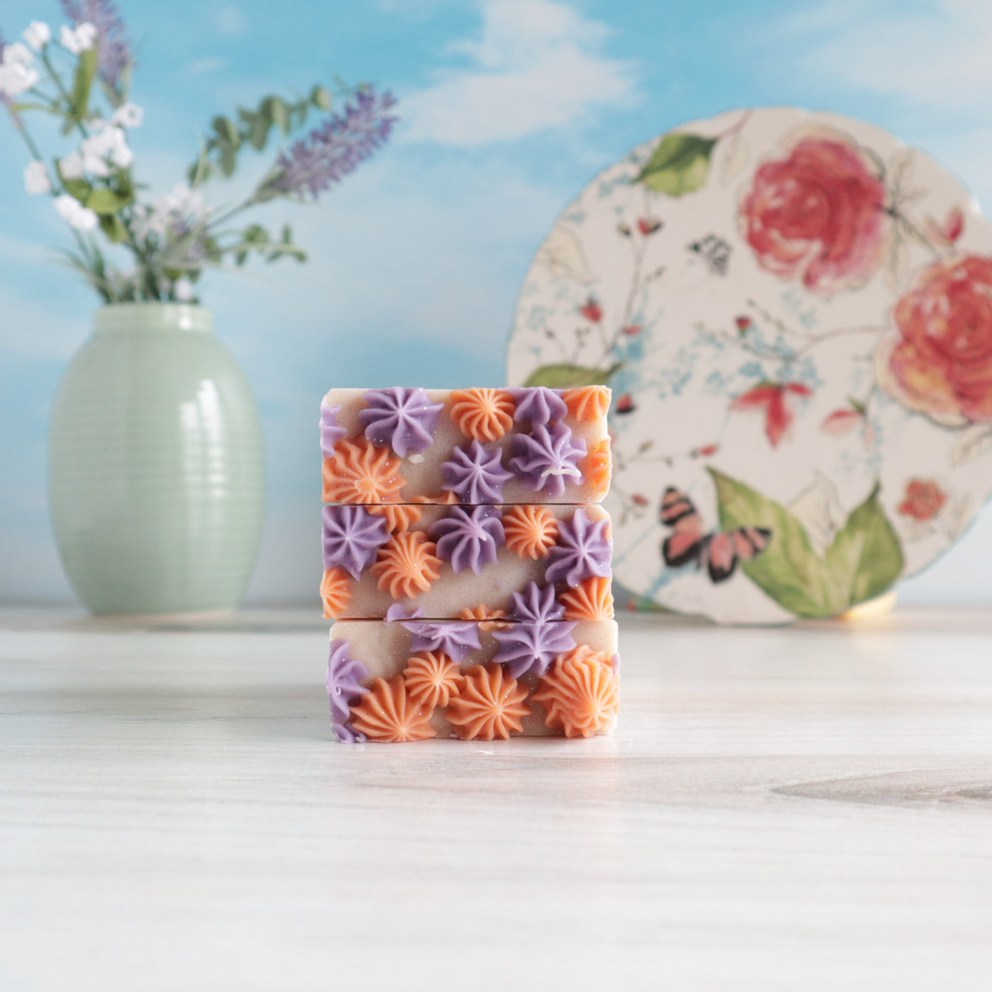 3 lavender apricot soaps are laying flat, stacked to show the pretty piping on top. there is a floral plate in the background standing of end to show the top and there is a vase of green with lavender buds in the back left of the image.