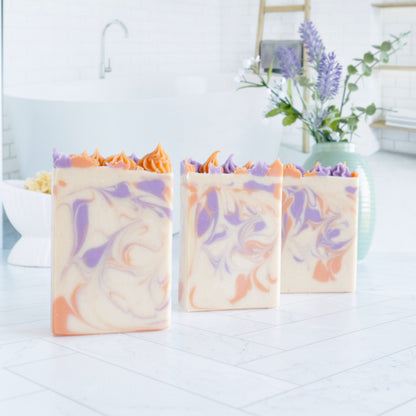 there are 3 lavender apricots standing in a line with the left closed to you, going back in a diagonal. they are showing the very pretty swirls of purple and orange in a creamy base, and you can get small glimpses of the tops with the piping peaking up. there is an image of a tub in the background and also there is a sage green vase in the back right with some lavender buds and greenery.