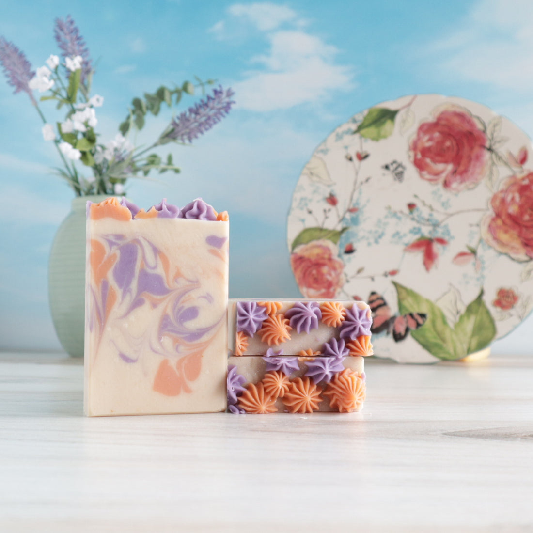 2 lavender apricot soaps are laying flat, stacked to show the pretty piping on top. there is a third one standing tall to show the muted colors of orange and purple in the creamy base color of the soap. It invokes a soothing vibe that is perfect pairing for the scent. there is a floral plate in the background standing of end to show the top and there is a vase of green with lavender buds in the back left of the image.