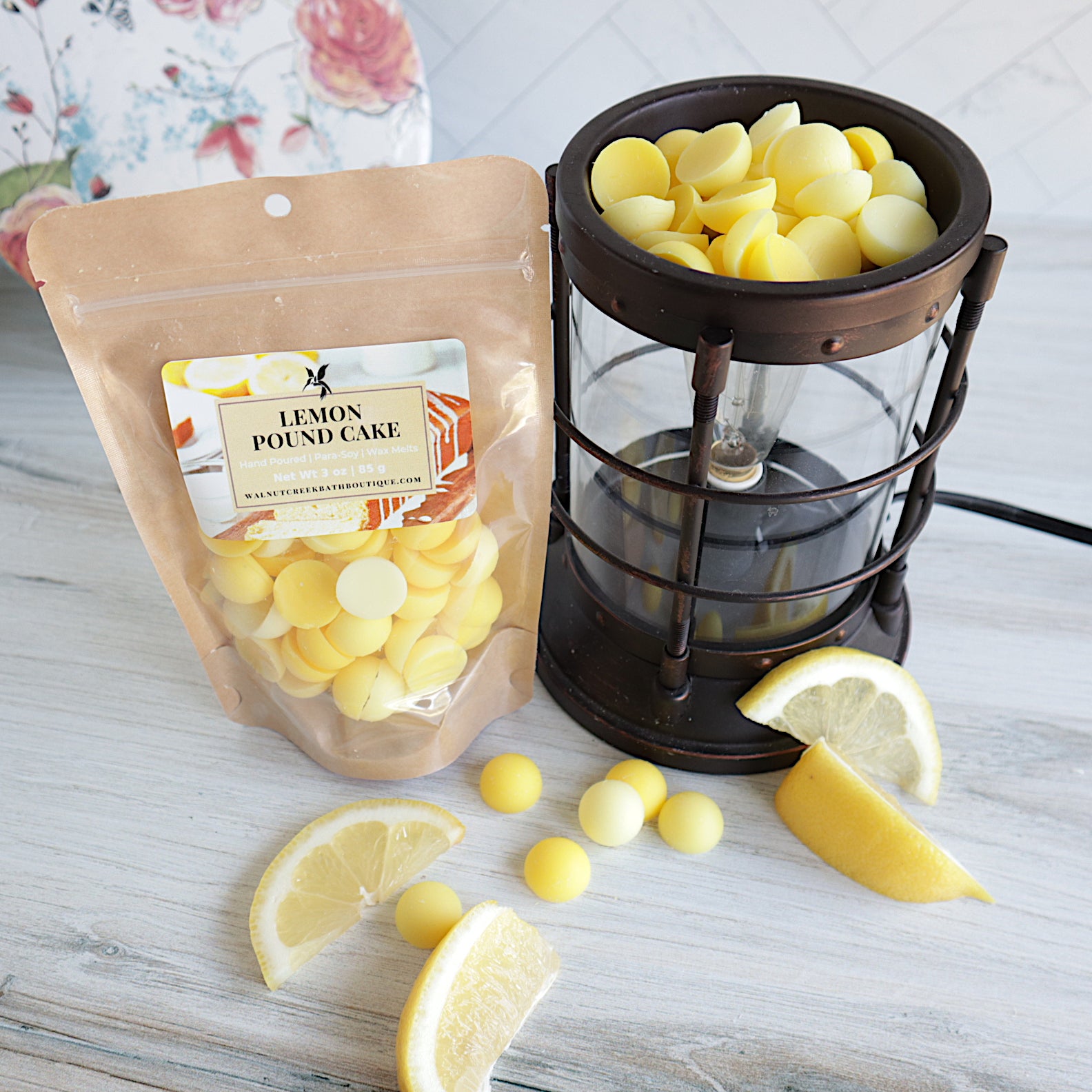 lemon pound cake wax melt bag is sitting next to a wax burner full of wax pieces. the wax are different shades of yellow. there are some wax pieces scattered around the base of the burner along with some fresh lemon pieces. there is a pretty floral plate in the background 