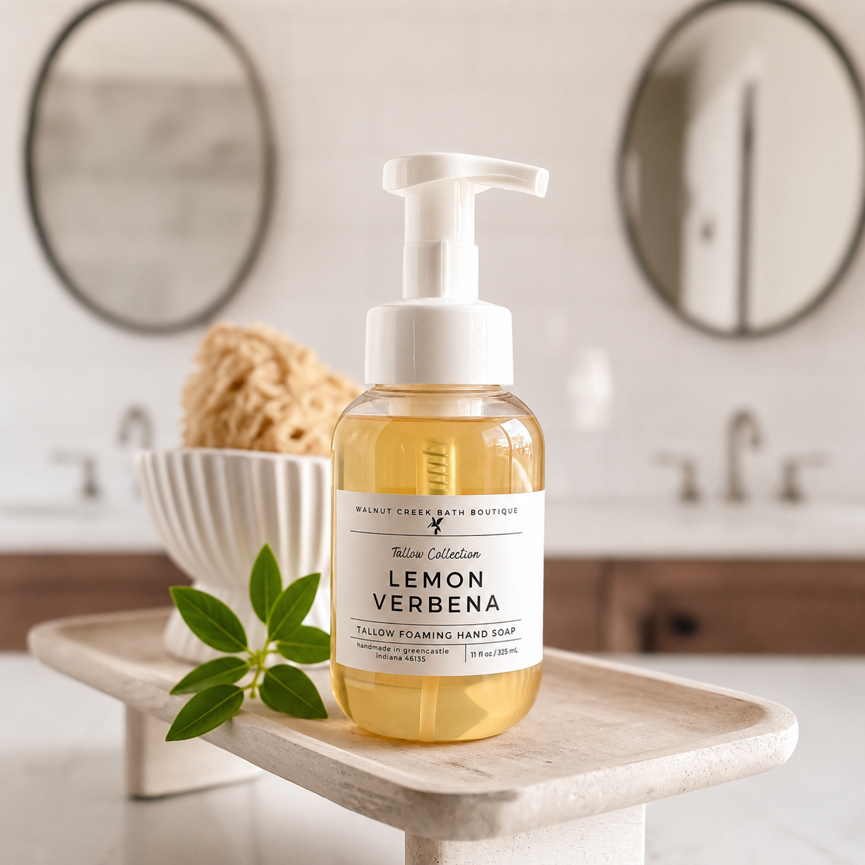 A bottle of Lemon Verbena yellow foaming hand soap by Malibu Clean Bath Boutique sits on a light stone tray in a bathroom setting. The clear bottle shows the golden-yellow liquid inside, with a white pump dispenser and a white label.
