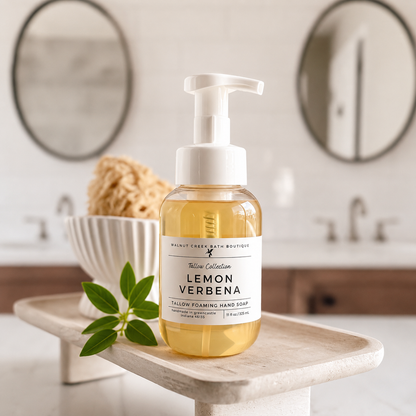 A bottle of Lemon Verbena yellow foaming hand soap by Malibu Clean Bath Boutique sits on a light stone tray in a bathroom setting. The clear bottle shows the golden-yellow liquid inside, with a white pump dispenser and a white label.