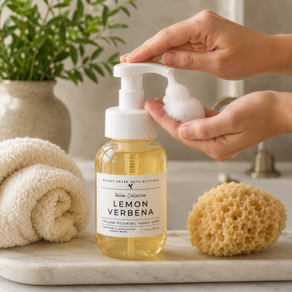 A bottle of Lemon Verbena hand soap with a white pump dispenser, being dispensed with foam in a bathroom setting. A folded beige towel, a natural sea sponge, and a potted plant are visible in the background.