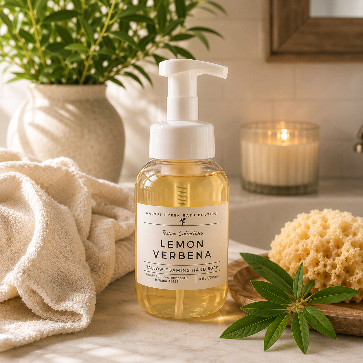 A bottle of Lemon Verbena Tallow Collection hand soap by Wainwright Creek Bath Boutique, styled on a marble countertop with a beige towel, a yellow sponge, a green leaf, and a lit candle in the background.