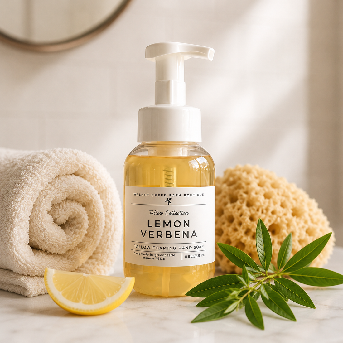 A bottle of Walnut Creek Bath Boutique Lemon Verbena Tallay Foaming Hand Soap stands on a marble surface with a rolled towel, lemon slice, sponge, and green leaves around it.