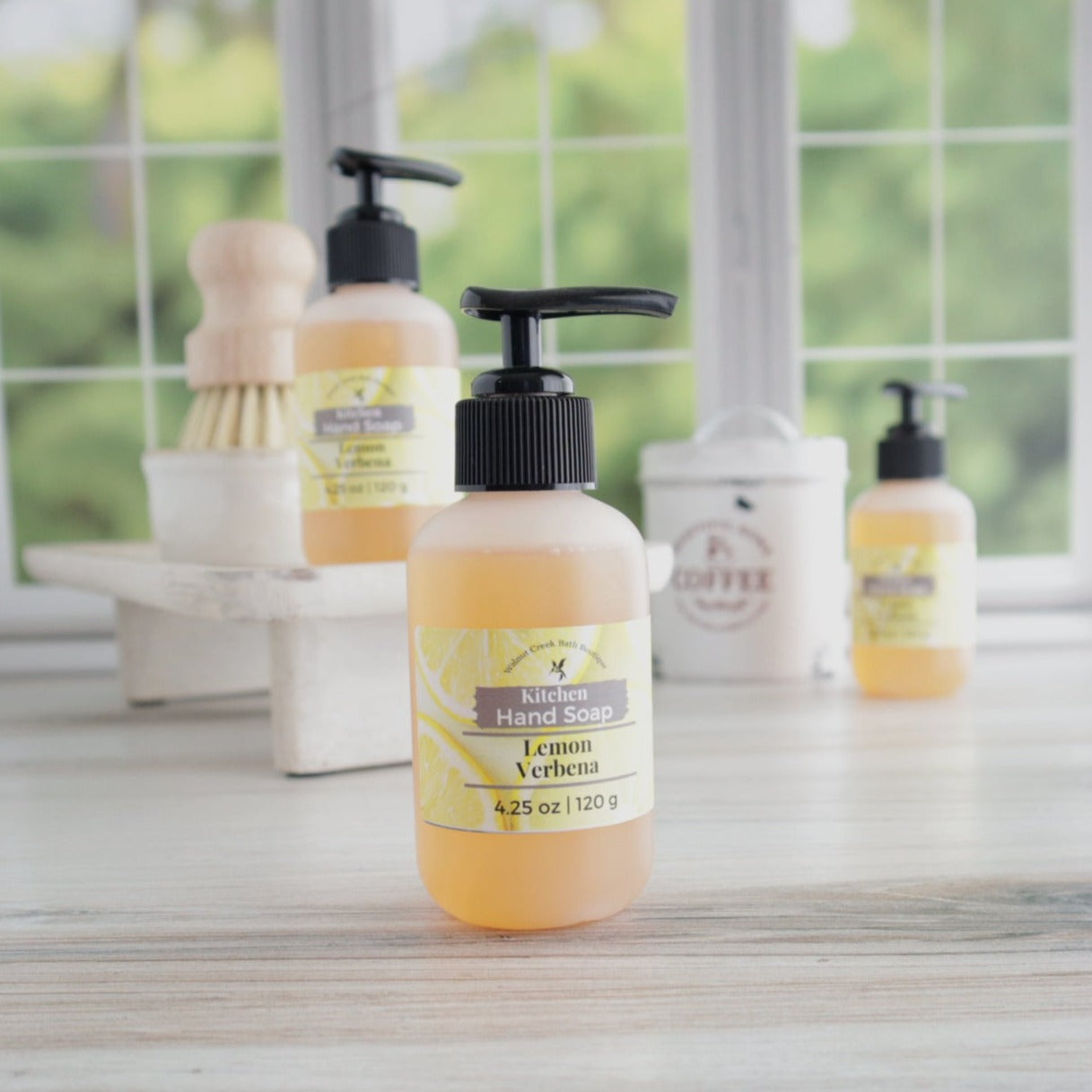 a lemon verbena hand soap is front and center in this picture. in the background is another one on a wooden tray that also is holding the solid dish soap and bamboo brush. in the very back and blurred out is a coffee tin and a third liquid soap. all of this is in front of a window with bright green trees in the background