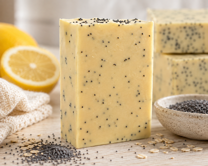 A rectangular bar of pale yellow soap speckled with black poppy seeds stands on a wooden surface, with lemon slices, oats, a small bowl of poppy seeds, and additional soap bars blurred in the background.
