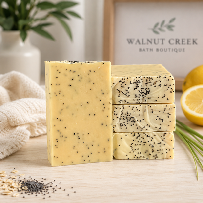 Three bars of pale yellow bath soap speckled with black poppy seeds, arranged on a light wood surface. A folded cream towel, a lemon slice, and a blurred background sign reading Walnut Creek Bath Boutique are visible.