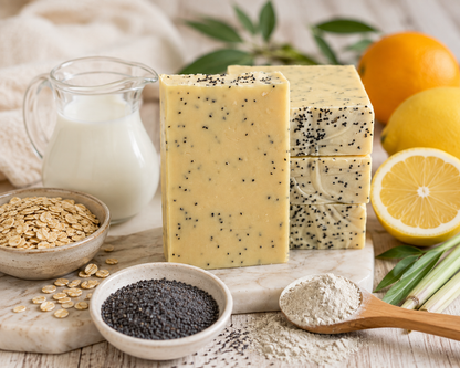 four rectangular bars of pale yellow soap speckled with black poppy seeds, displayed on a marble board with oats, milk, poppy seeds, flour, lemongrass, and citrus slices in a styled product setup.