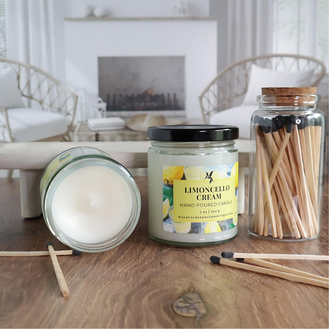 2 jars of limoncello cream candles are shown, one with the lid off and laying flat to show the smooth top and the other with the lid on and label facing you to show that. there are matches in a clear jar to the right of the candles and a few matches scattered around the table  as well. in the background is a fireplace flanked by 2 wicker chairs