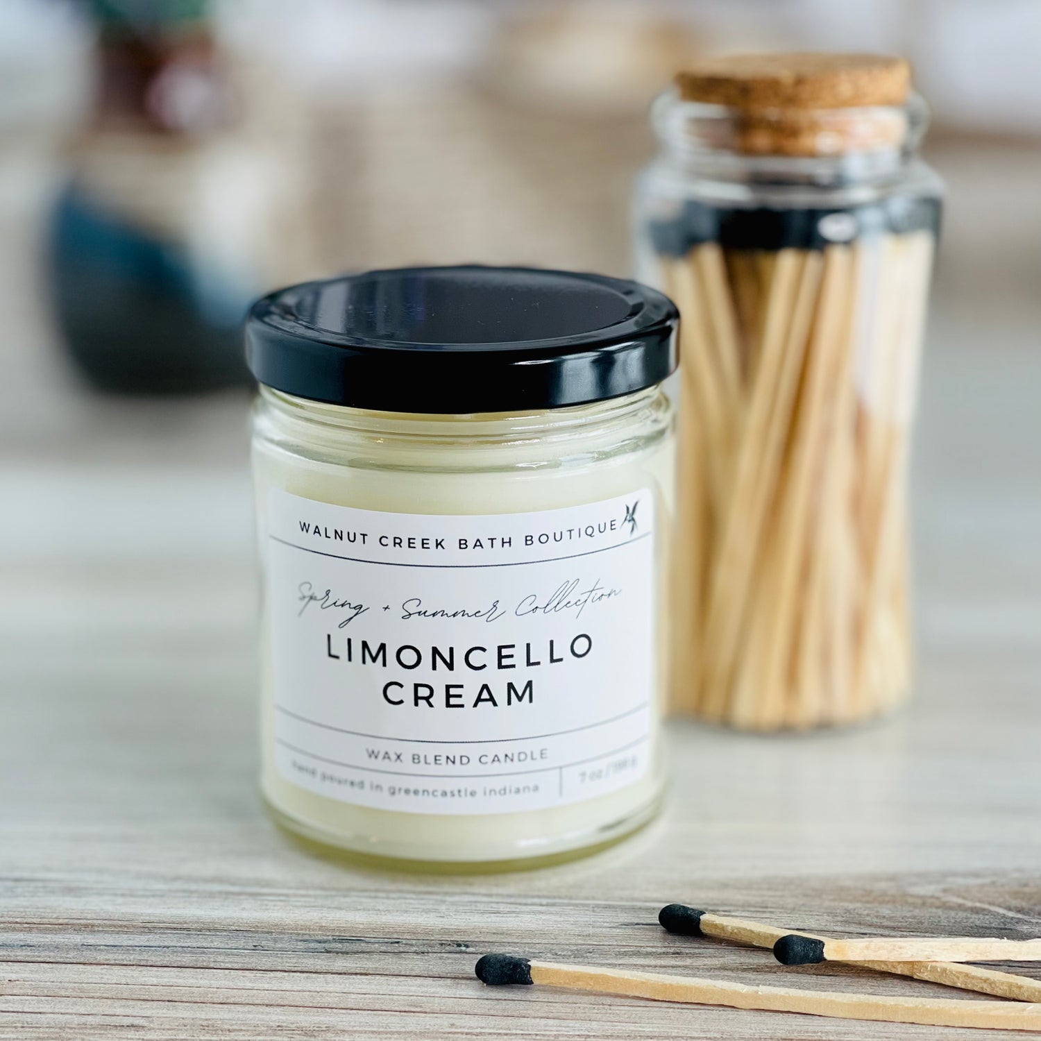 A Limoncello Cream candle in a clear glass jar with a black lid, labeled with the brand name and product title. The candle is placed on a wooden surface with matches and matchsticks in the background.