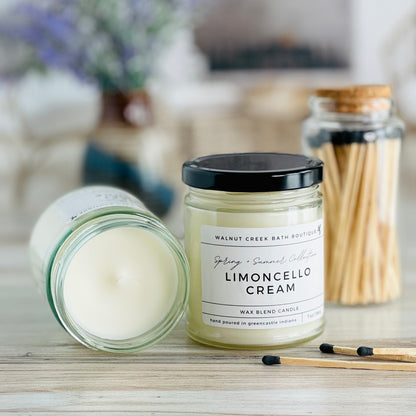 limoncello cream candle with lid to the right of another candle with the top off to show the creamy wax inside. there are a few matches scattered in front and a pretty glass jar of matches in the background