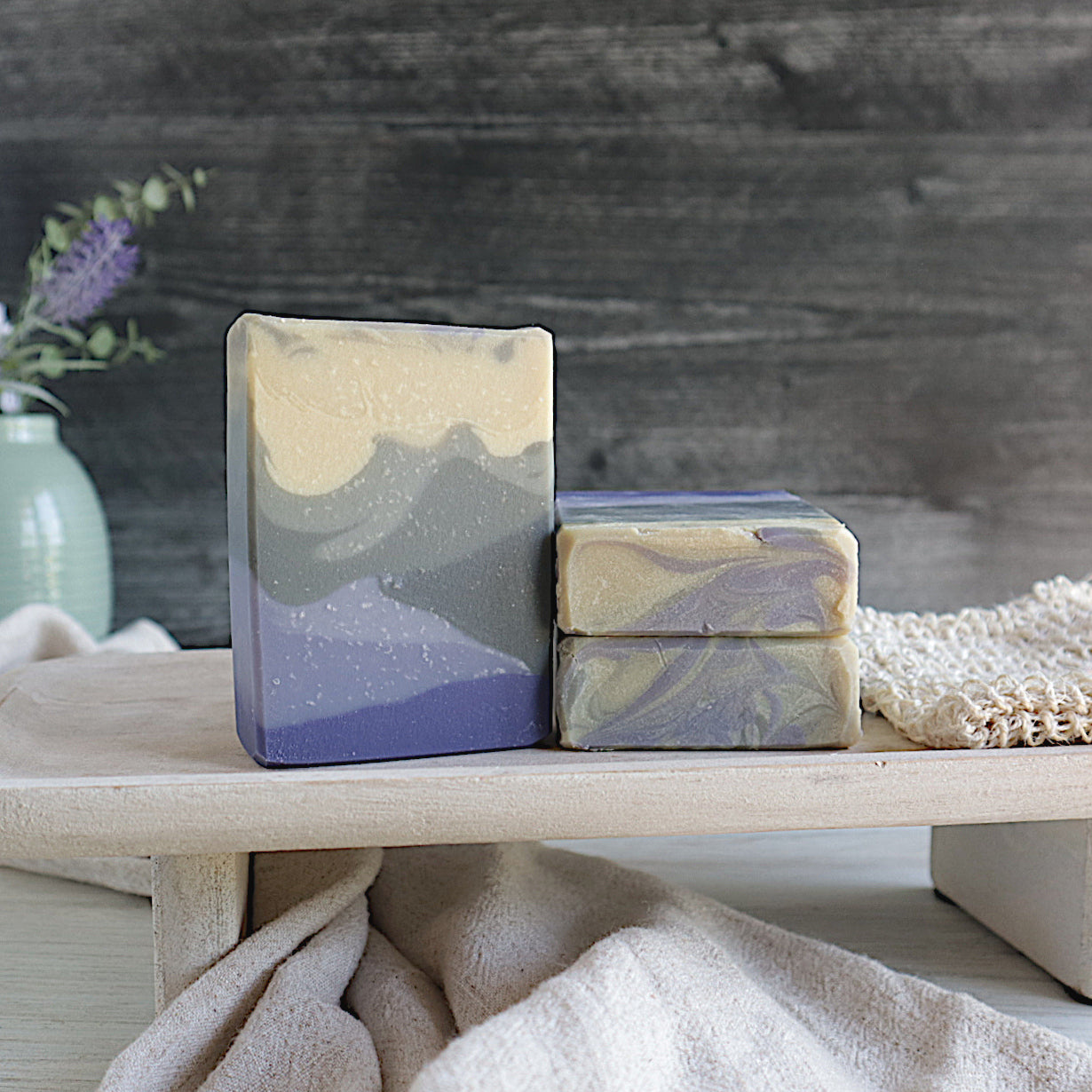 3 London fog soaps are shown sitting on a raised tray. one soap is standing tall facing you showing the purple bottle that gets lighter as it goes up into the grey middle that also gets lighter as it goes up into the creamy off-white top that has a small amount of grey swirls. next to these is a tan soap saver and underneath the tray is a cream colored towel running diagonal. the background is a black old wood wall and there is a green vase with lavender buds in the back left