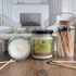 2 jars of merlins forest candles are shown, one with the lid off and laying flat to show the smooth top and the other with the lid on and label facing you to show that. there are matches in a clear jar to the right of the candles and a few matches scattered around the table as well. in the background is a fireplace flanked by 2 wicker chairs