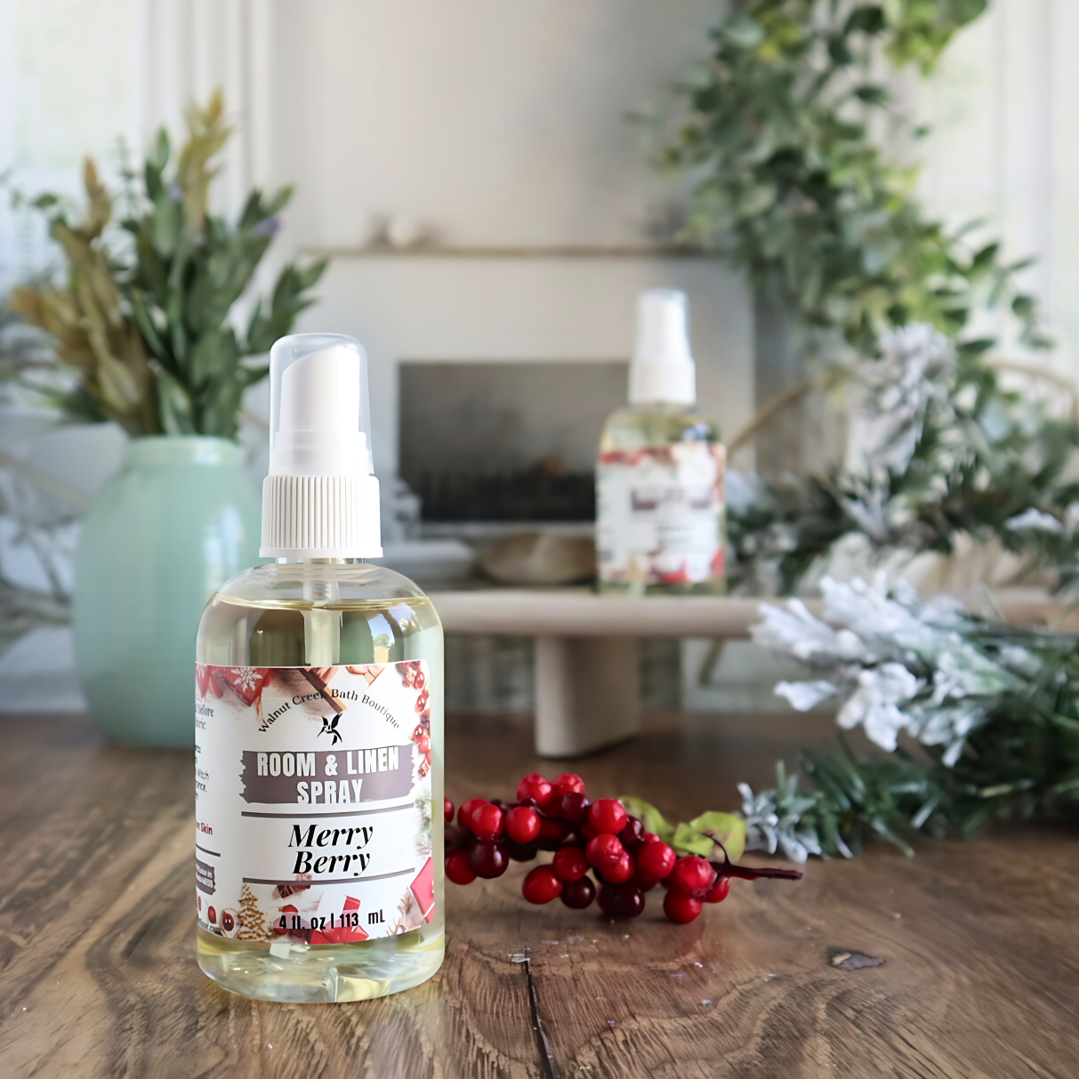 merry berry room spray is shown. in the background is a second bottle on a tray next to a green vase with small flower buds. there are snow covered pine branches surrounding the back bottle and some red berries on a stem behind the front bottle.  this is in front an image of a fireplace flanked by two chairs. there is some greenery hanging from the back right.