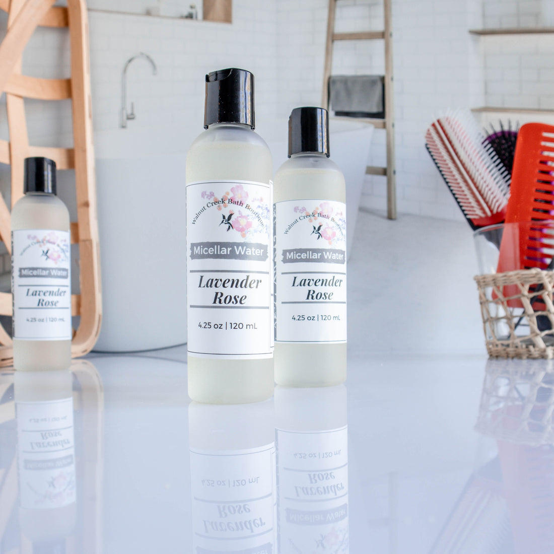 3 lavender rose micellar waters are shown, 2 closer to viewer and a third in the back left. there is a bathroom scene in that background showing a tub. there is also a  cup with hair brushes and combs in the back right.