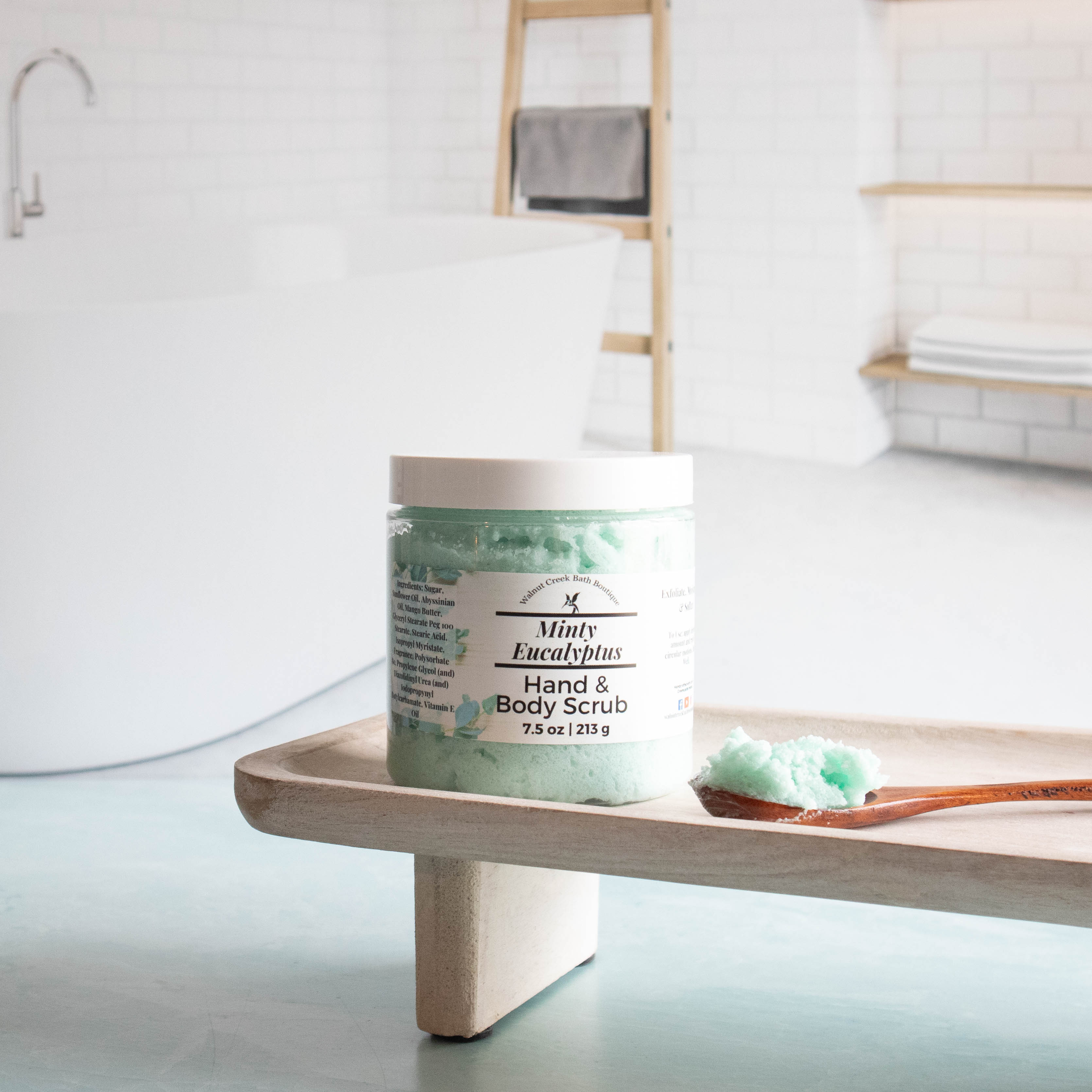 minty eucalyptus scrub is sitting on a tray. there is a wooden spoon filled with some scrub laying on the try next to the jar. this shows the pretty minty green scrub color. there is a tub in the background with a ladder with a towel draped over one of the rungs.