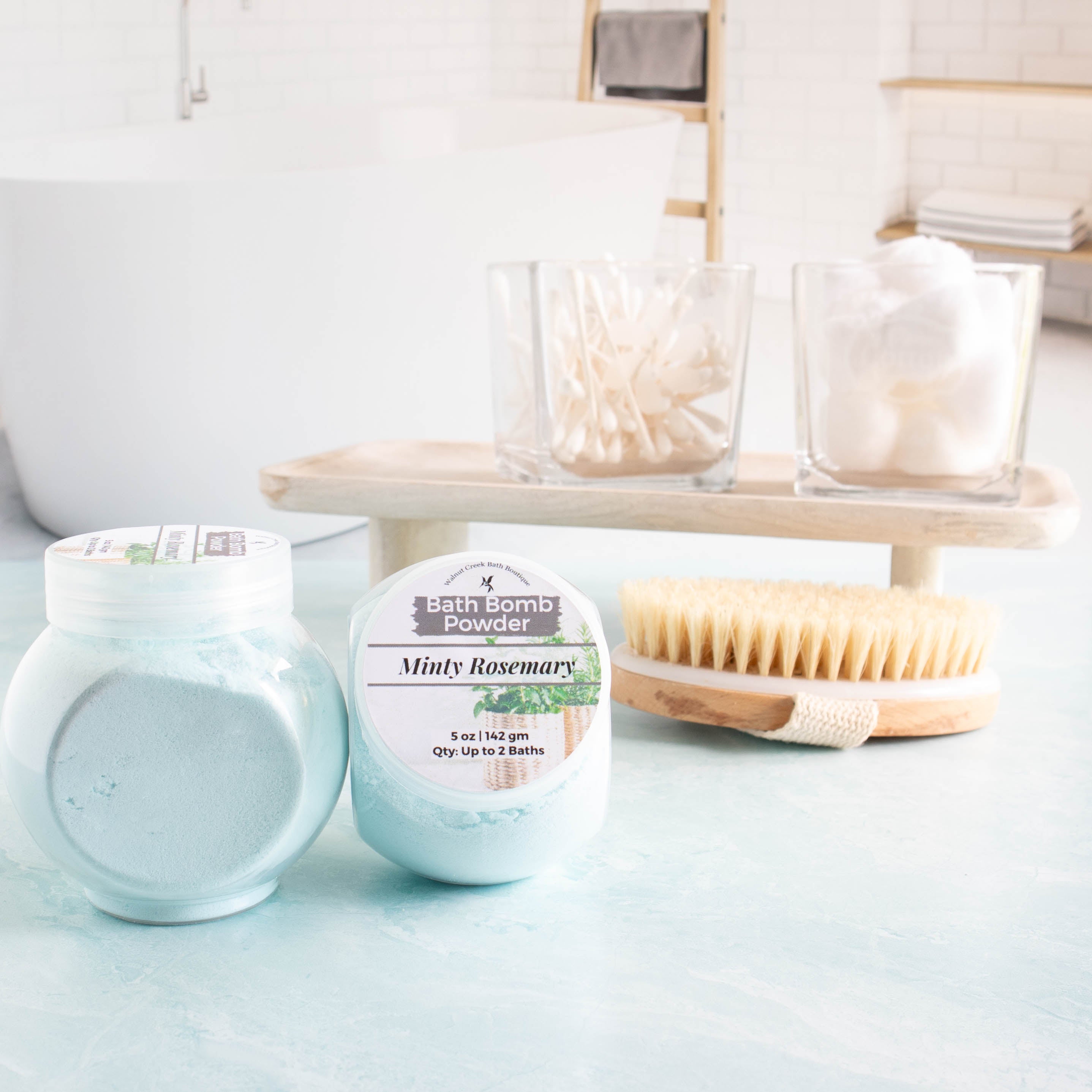 two 5 oz jars of blue minty rosemary bath bomb powder in the foreground. One showing the label and another showing the side of the jar with the powder.In the background is a tray with cotton balls and q-tips and a body brush. there is a spa like bath room in the background