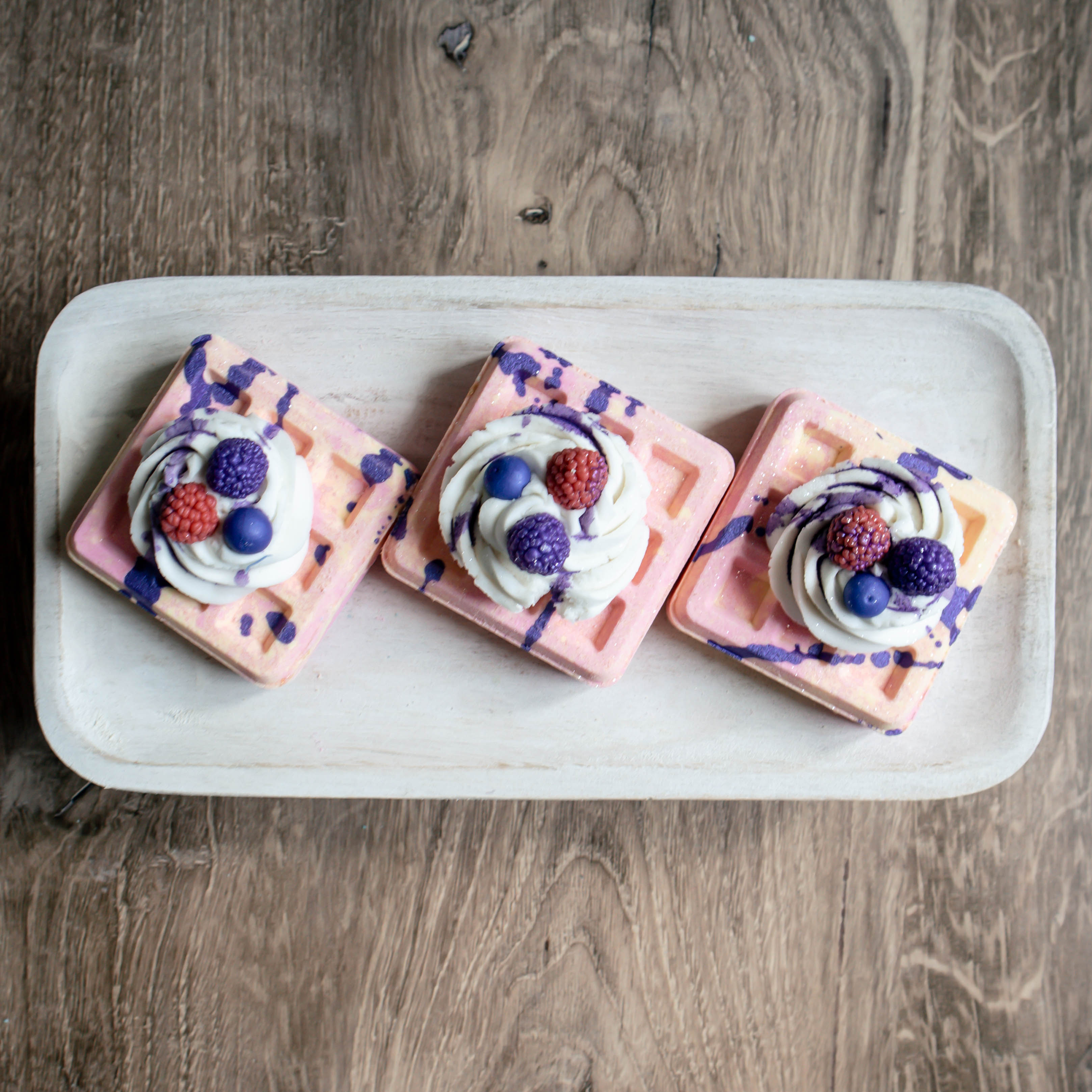 3 waffle bath bombs sitting on a tray. these waffles are pink and yellow and have a huge dollop of bubble bath frosting. On top of the frosting is some soapy berries and then there is a purple drizzle over all of it, just to make it extra fun!