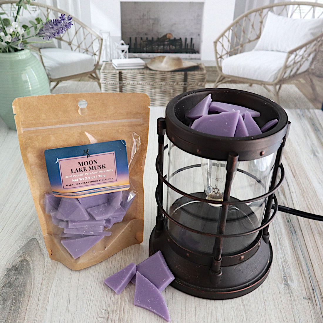 moon lake musk wax melts are in a bag that is standing next to a wax burner. the wax melts are purple in color and shards in shape. the burner is full of these shards while there are some that are piled on the table in front of it.  this is all sitting on a washed out wooden base with a couple of chairs in the background next to a fireplace