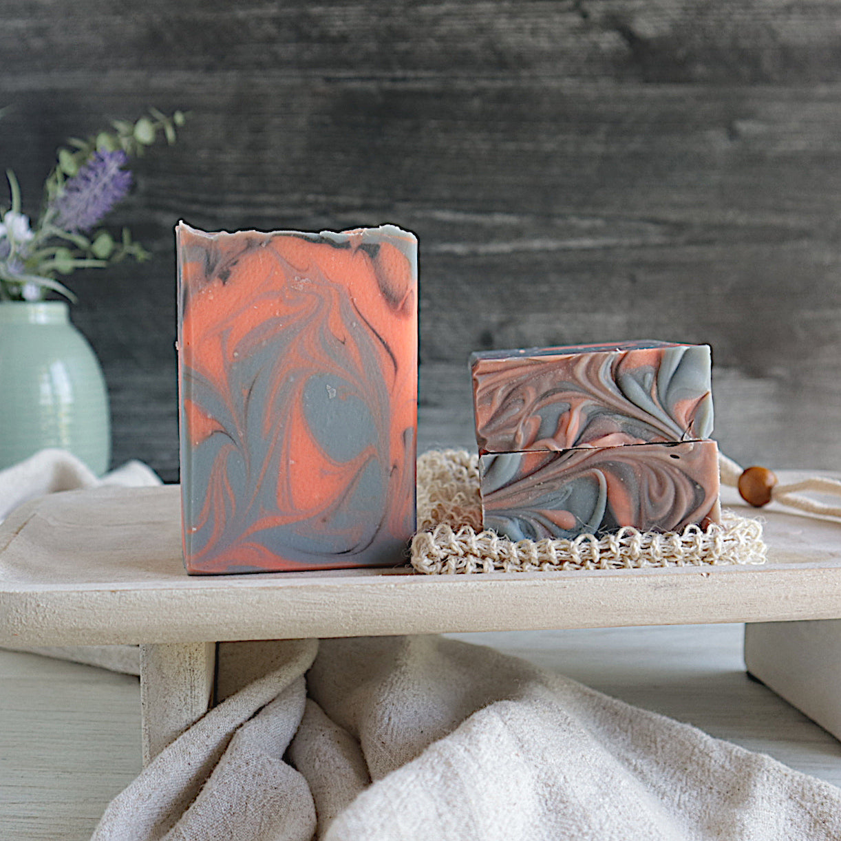 morning dew is shown with one bar to the left sitting on a try. next to this are 2 more bars laying flat on a soap loofah, the tops are forward to show the pretty swirled top. the soap is orange with blue and a small amount of black swirls. in the background is a sage green vase against a black wood backdrop