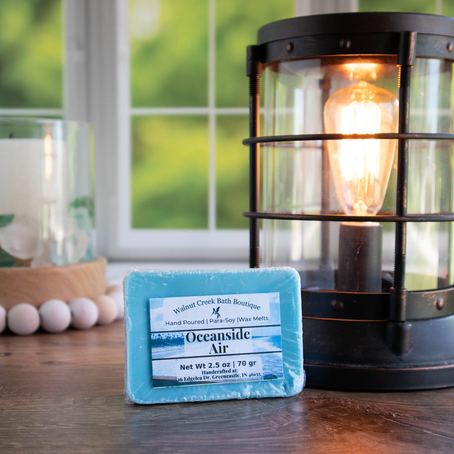 Oceanside Air is pictured. It is a pretty blue color. Laying up against a lit wax warmers. There is a candle with a wood bead in the background and in the very back view is a window with some green leaves showing.