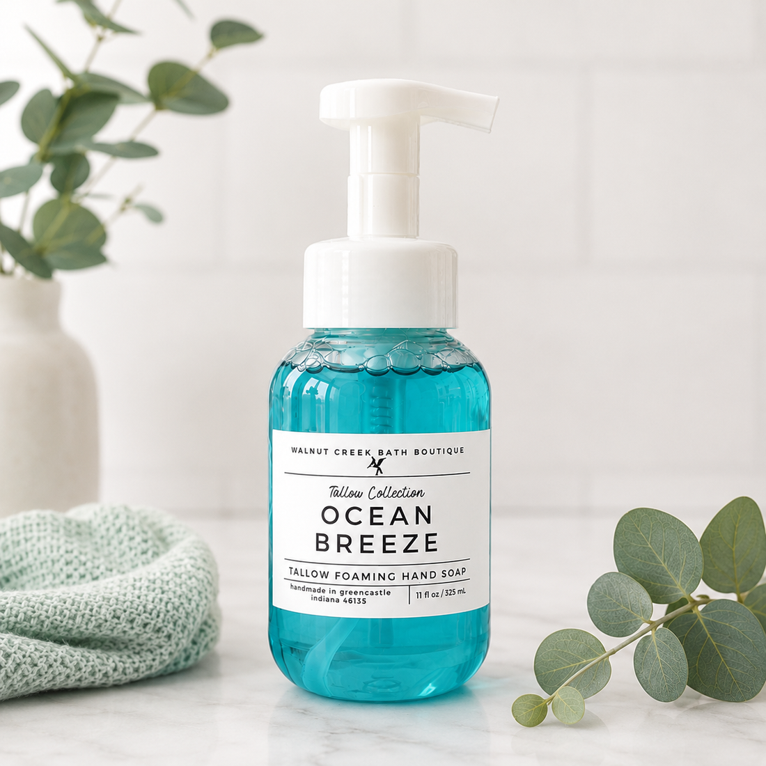 A bottle of Ocean Breeze Fallow Foaming Hand Soap by Walnut Creek Bath Boutique, featuring a translucent blue bottle and a white pump dispenser, styled on a marble surface with eucalyptus leaves and a mint green towel.