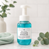 A bottle of Ocean Breeze Fallow Foaming Hand Soap by Walnut Creek Bath Boutique, featuring a translucent blue bottle and a white pump dispenser, styled on a marble surface with eucalyptus leaves and a mint green towel.