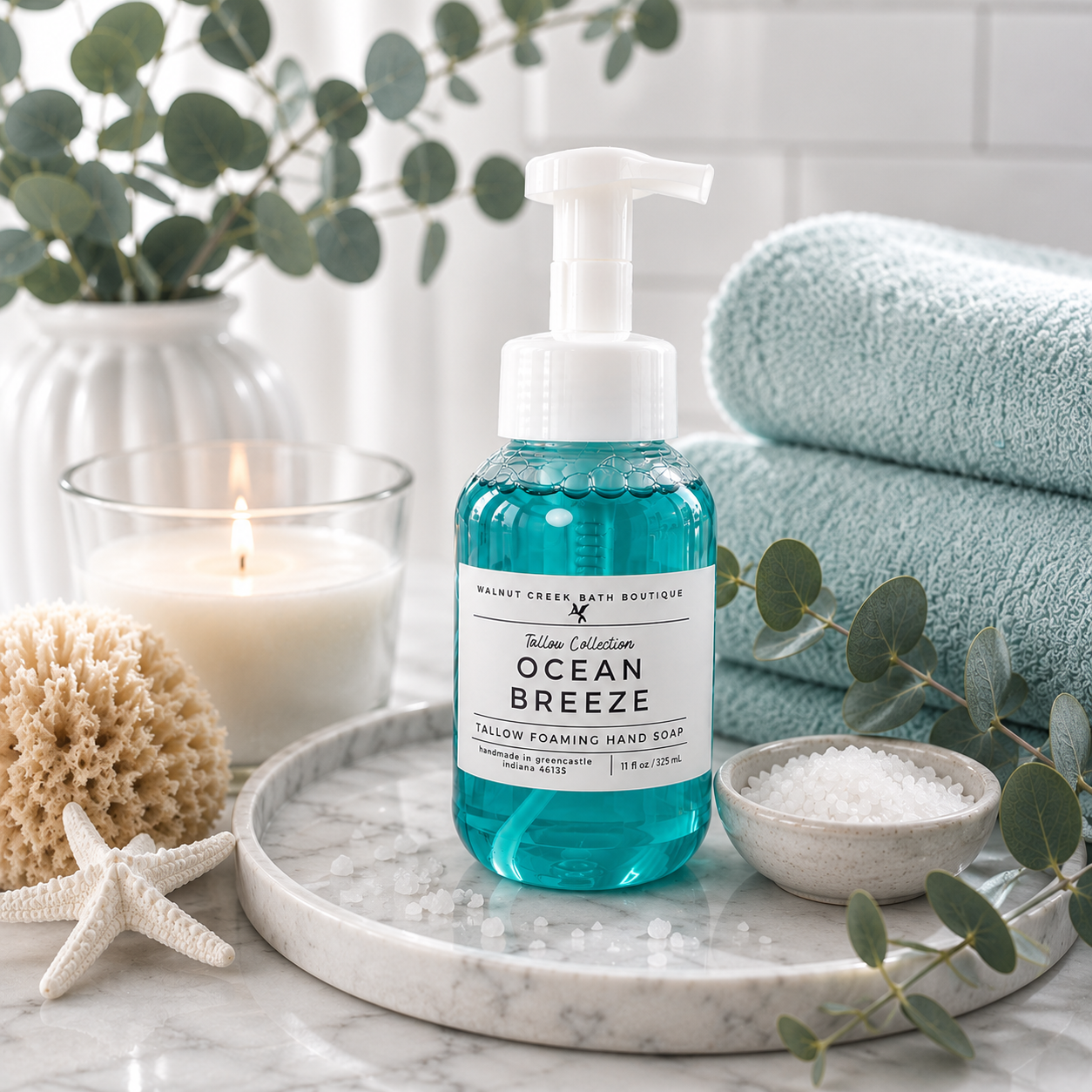 Ocean Breeze Salty Foaming Hand Soap by Malibu Creek Bath Boutique in a clear bottle with turquoise liquid, styled on a marble tray with a candle, eucalyptus, towels, and bath salts.